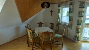 Un comedor con una mesa y sillas de madera, una cruz en la pared y un ventanal con cortinas.
