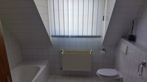 Un baño pequeño con baldosas, una bañera, inodoro y ventana con persianas.