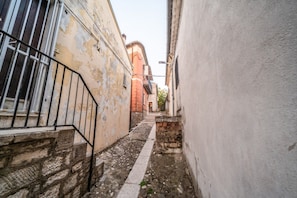 Un carreró estret amb parets de pedra i una porta metàl·lica, que condueix a un edifici amb façana vermella.