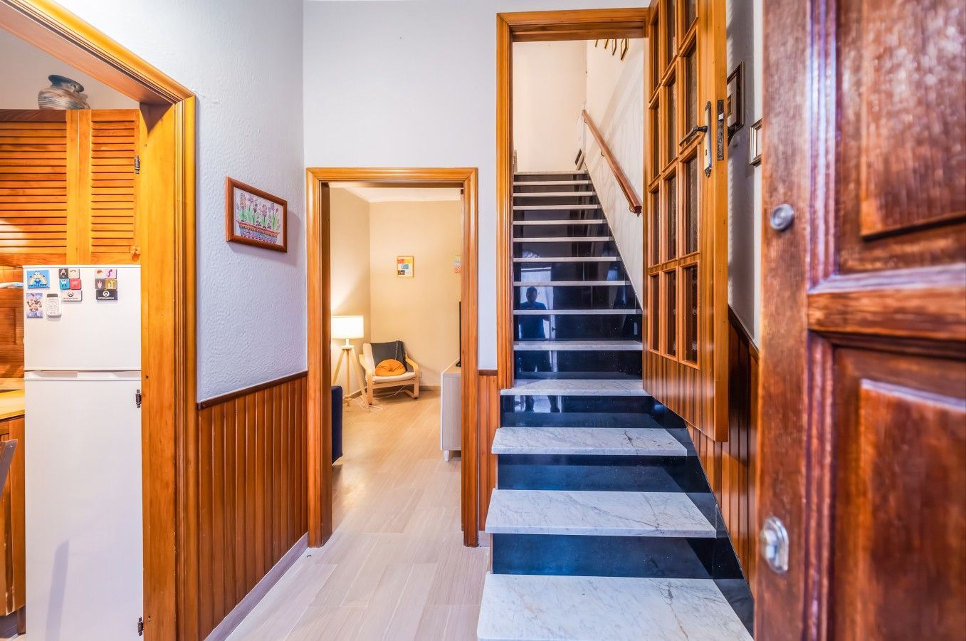 Un couloir avec une porte en bois, un réfrigérateur orné d’aimants, une chaise et un escalier avec une rampe en bois.