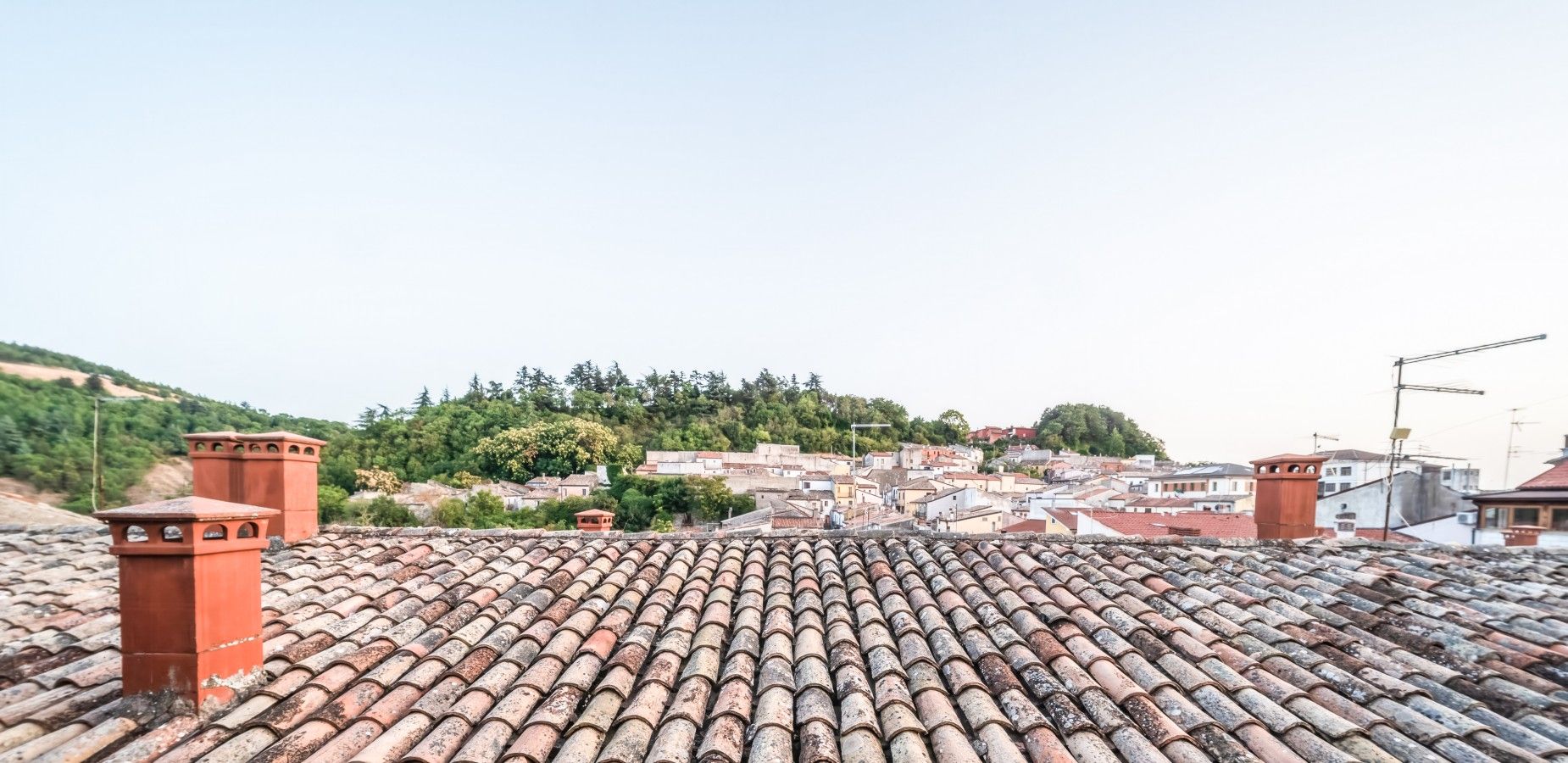 A rooftop with terracotta tiles, a view of a town with buildings, and a hill with greenery in the background.