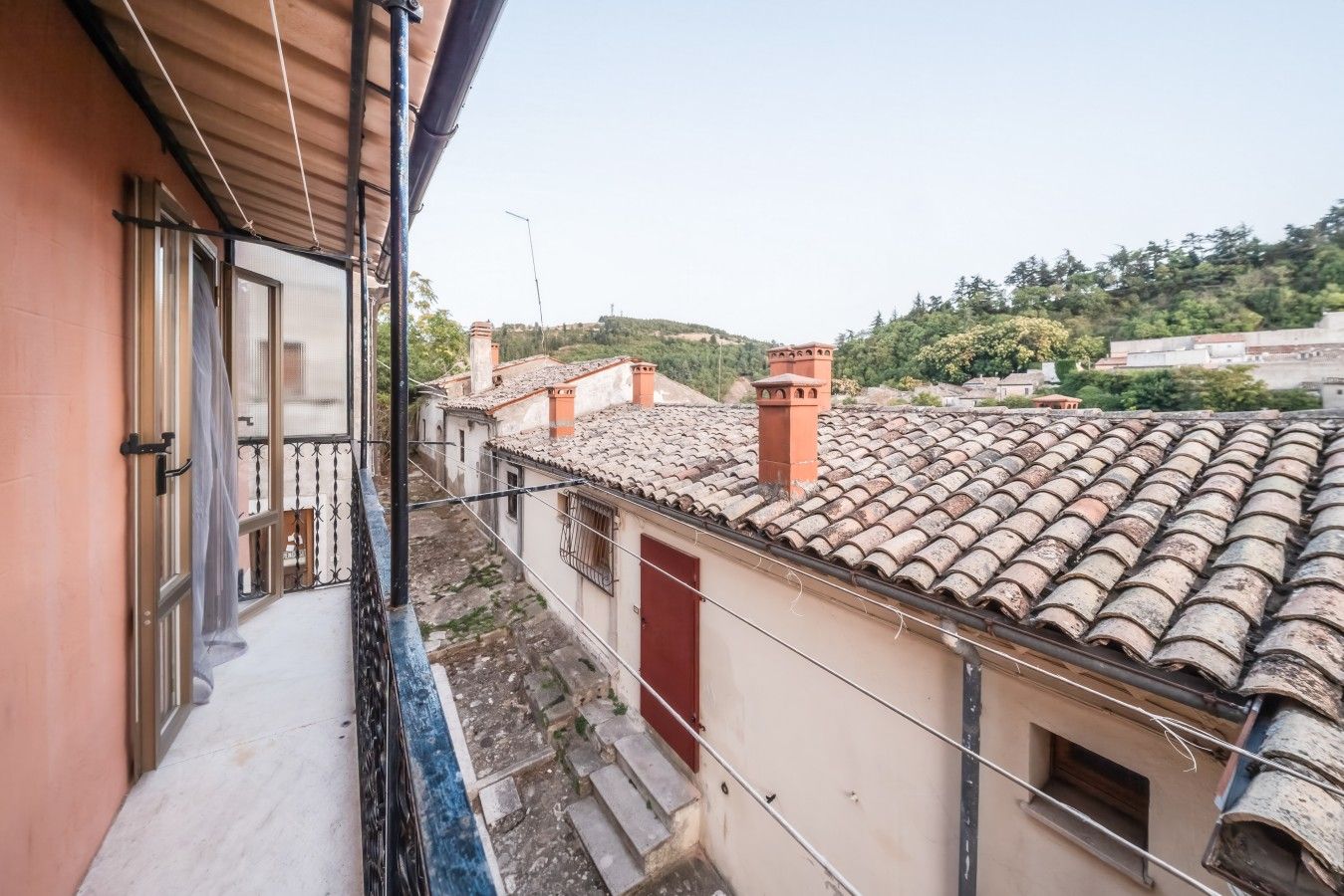 Un balcon étroit avec une rambarde en métal, une porte rouge et une vue sur les bâtiments voisins, dont les toits sont recouverts de tuiles en terre cuite.