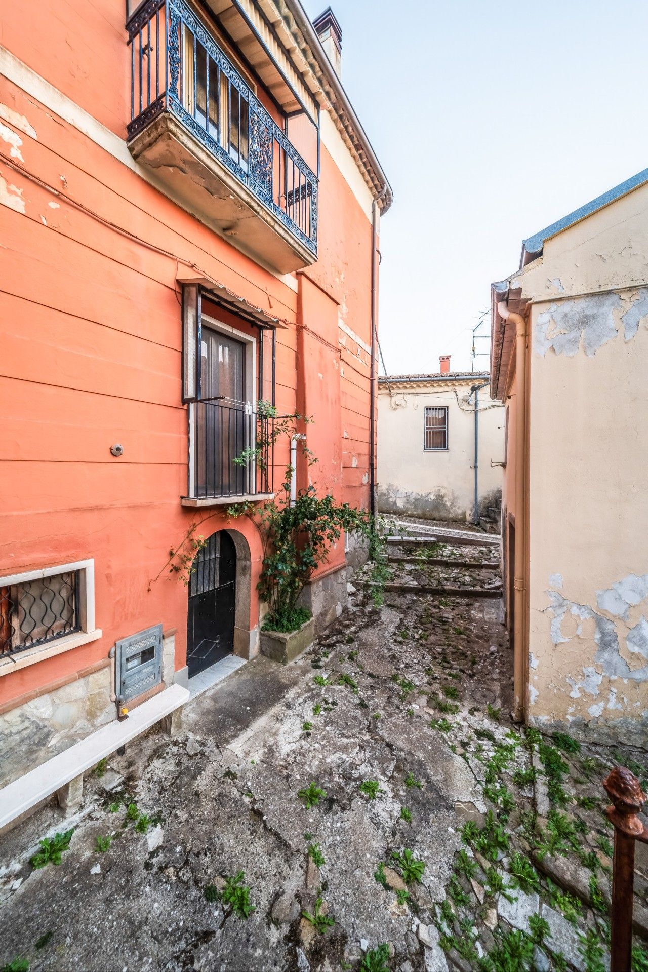 Une ruelle étroite, avec un immeuble sur la gauche qui présente une porte noire et une fenêtre ornée d’une grille métallique.