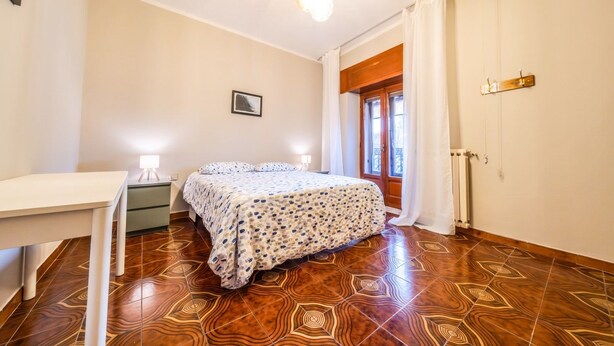 Une chambre à coucher comprenant un lit, des tables de chevet, un bureau, un radiateur et une fenêtre avec des rideaux.