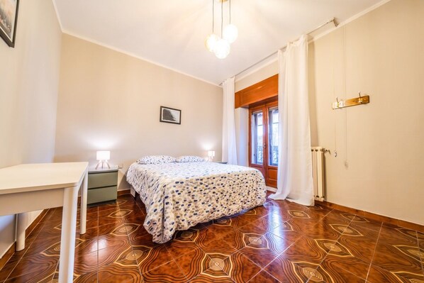 A bedroom with a bed, bedside tables, a desk, a radiator, and a window with curtains.