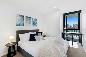 A hotel room with a bed, two bedside tables, a window with a city view, and abstract wall art. - Ocean View 2BR, Steps to Beach | Pool, Gym Parking (Broadbeach)
