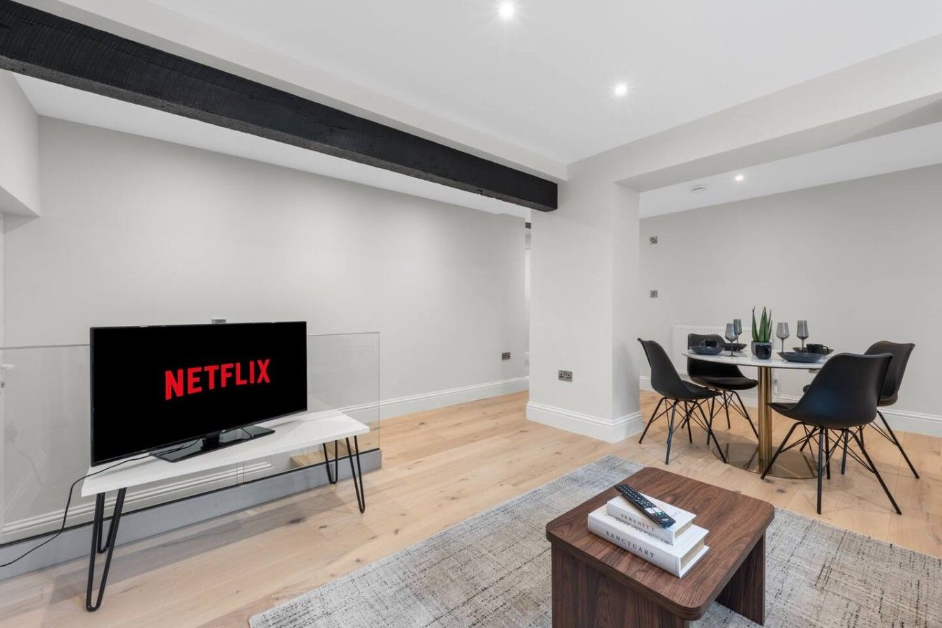 A modern living room with a flat-screen TV displaying 'NETFLIX,' a dining area with a table and chairs, and a small wooden coffee table with books.