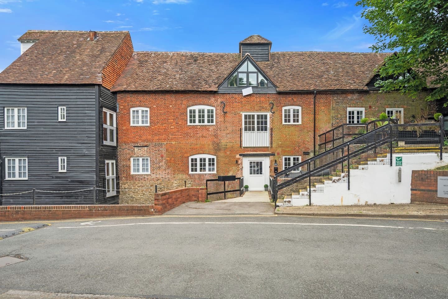 A two-story building with a brick facade, a staircase leading to the entrance, and a gated driveway.