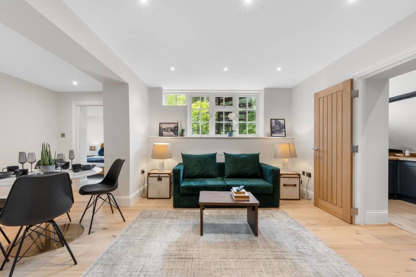 A modern living room with a green sofa, a coffee table, and a dining area with chairs and a table. There is a window with a view of greenery and a door leading to another room.