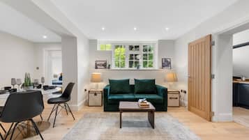 A modern living room with a green sofa, a coffee table, and a dining area with chairs and a table. There is a window with a view of greenery and a door leading to another room.