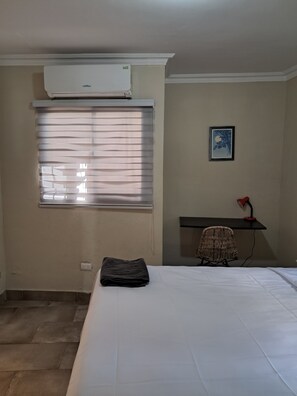 Habitación de hotel con cama, escritorio, silla, ventana con persianas y aire acondicionado.
