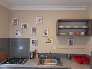 Cocina con estufa a gas, lavamanos y diversos utensilios de cocina. Hay cuadros enmarcados en la pared y una estantería con vajilla.