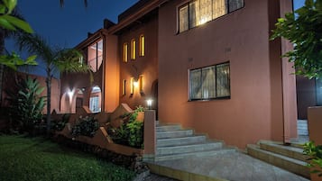 A two-story building with a staircase leading to the entrance, surrounded by greenery and palm trees.