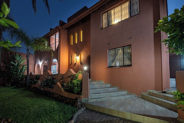 A two-story building with a staircase leading to the entrance, surrounded by greenery and palm trees.