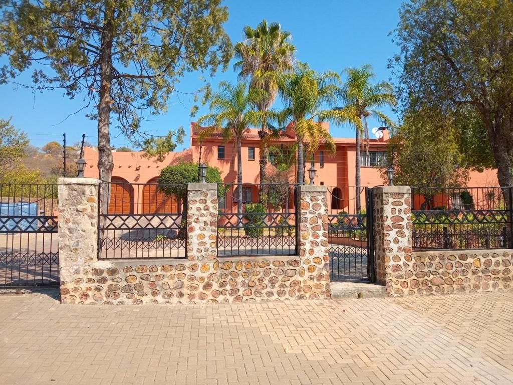 A stone wall with metal gates, a paved walkway, and a building with a red facade and palm trees in the background.