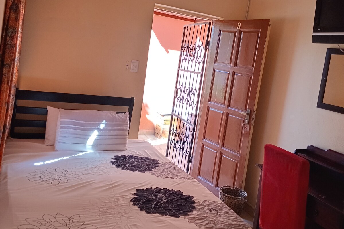 A bedroom with a bed, a ceiling fan, an air conditioning unit, a red chair, and a TV on the wall.