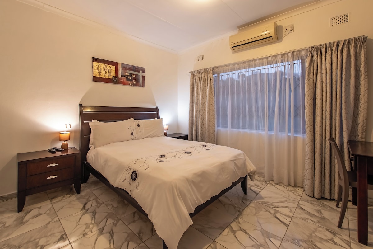 Habitació d'hotel amb un llit, taules de nit, una làmpada, un aparell d'aire condicionat i una finestra amb cortines.