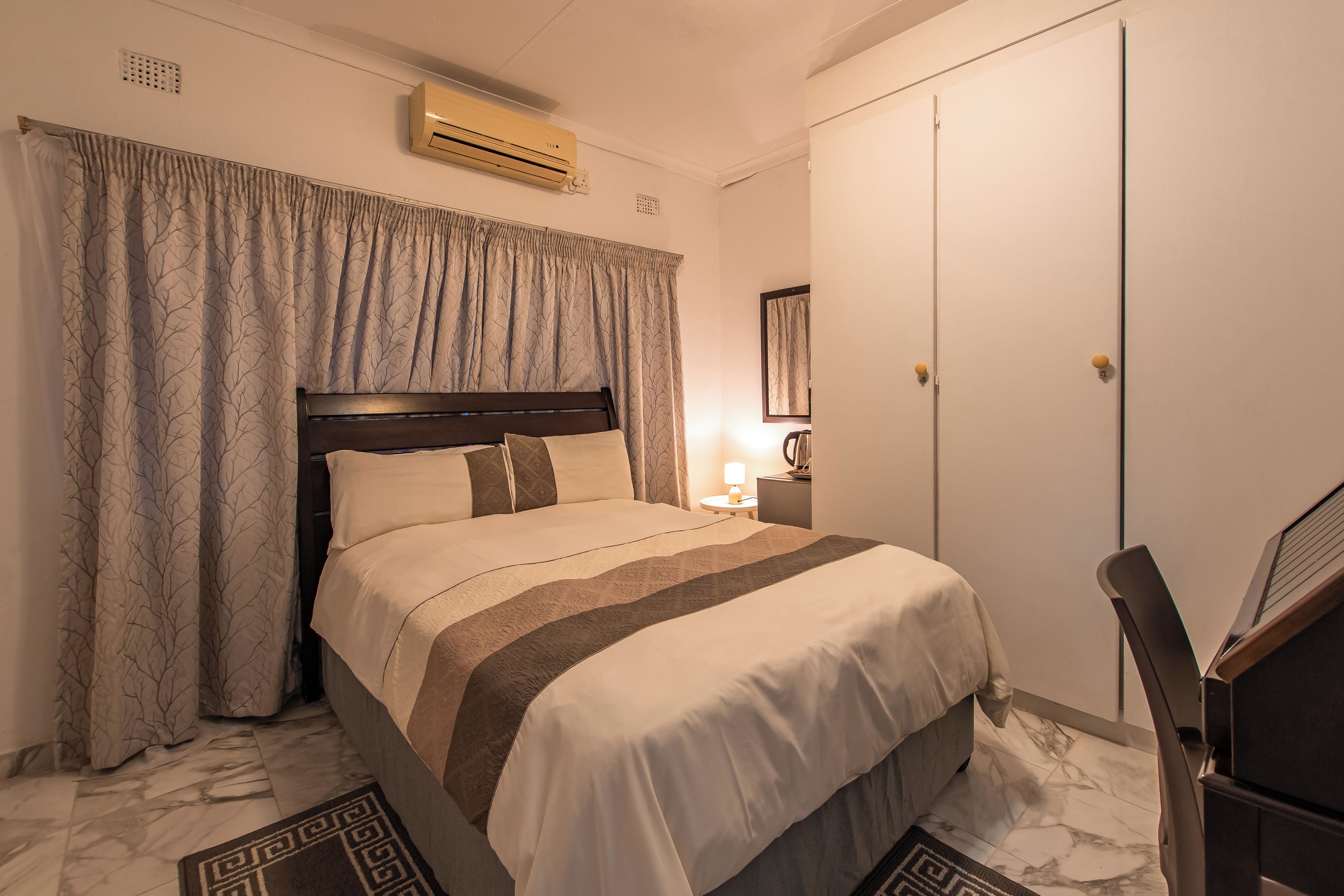 A hotel room with a bed, curtains, a bedside lamp, a wardrobe, and an air conditioning unit.