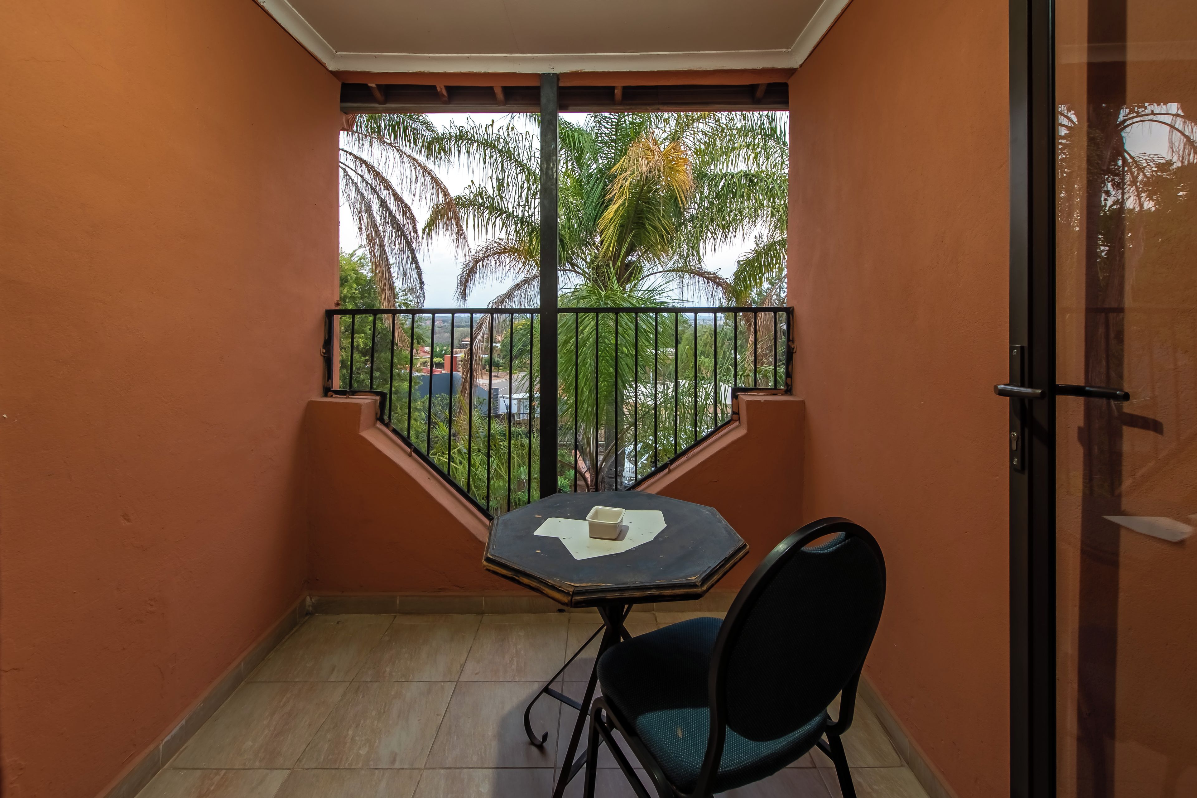 A small balcony with a table and chair, a view of palm trees and buildings, and a tiled floor.