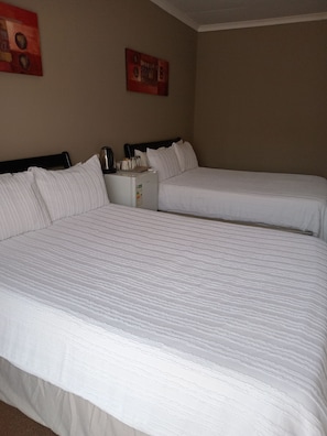 Quarto de hotel com duas camas, uma mesinha com uma chaleira e um relógio de parede.