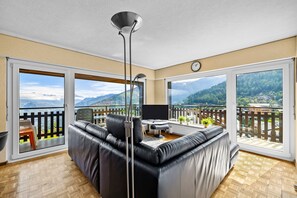Ein modernes Wohnzimmer mit einer schwarzen Ledersofa, einem Flachbildfernseher und einem Essbereich mit Tisch und Stühlen. Große Fenster bieten Blick auf eine gebirgige Landschaft.