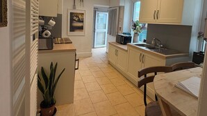 A compact kitchen with white cabinets, a dining area with a wooden table, and a potted plant. - Bude Surf Shack | Walk to the beach | Dog-friendly | Sleeps 5 (Bude)