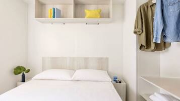 A bedroom with a bed, a bedside table, a shelf with books and a pillow, and a clothing rack.