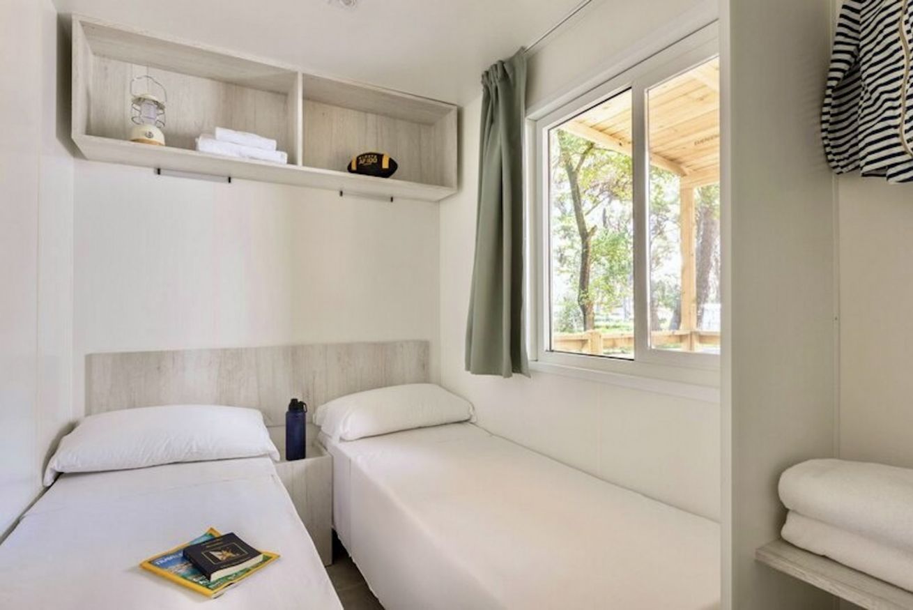 Camera da letto con due letti, uno scaffale sopra i letti, una finestra con tende e vista su alberi.