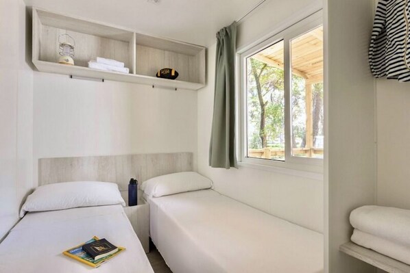 A bedroom with two beds, a shelf above the beds, a window with curtains, and a view of trees outside.