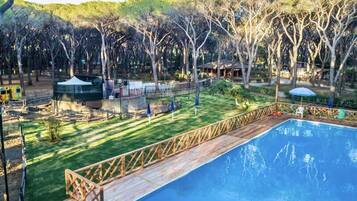 Una piscina con plataforma de madera, rodeada de césped, árboles y un parque infantil vallado.