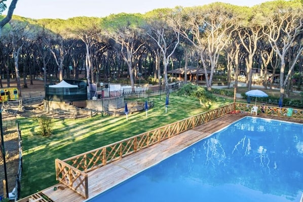 A swimming pool with a wooden deck, surrounded by a grassy area with trees and a fenced playground.