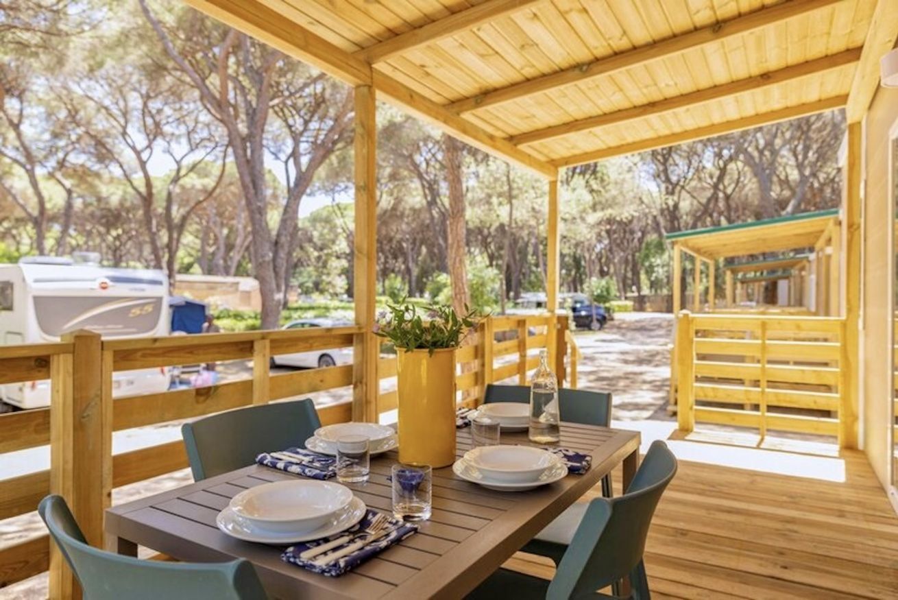 An outdoor dining area with a wooden table set for two, surrounded by chairs, and a covered seating area with a view of the surrounding trees.