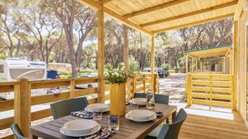 An outdoor dining area with a wooden table set for two, surrounded by chairs, and a covered seating area with a view of the surrounding trees.