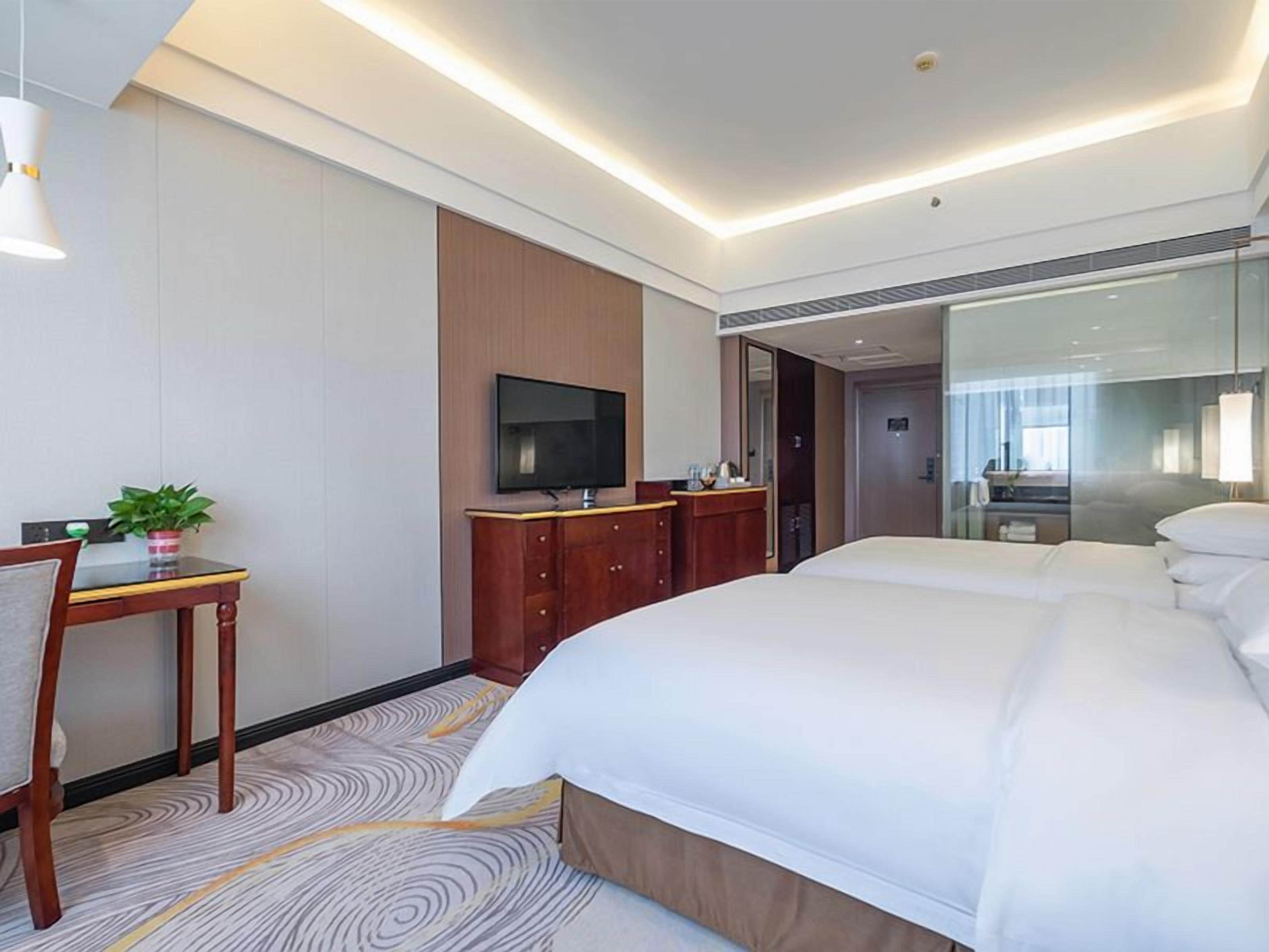 A hotel room with a large bed, a television, a desk with a plant, and a mirror.