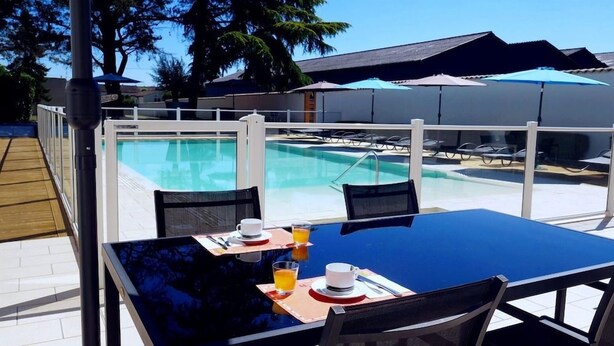 An outdoor pool area with a table set for two, a chair, and a sun umbrella. - Hotel Le Capricorne Rest. Le Trianon (Marmande)