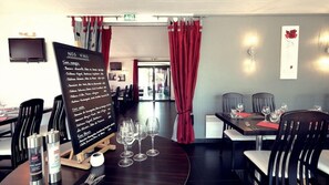 A restaurant interior with a menu board, dining tables, chairs, and a television mounted on the wall. - Hotel Le Capricorne Rest. Le Trianon (Marmande)