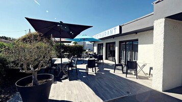 Un espacio de patio al aire libre con mesas y sillas, un árbol en maceta y un edificio con la leyenda 'RESTAURANT' al fondo.