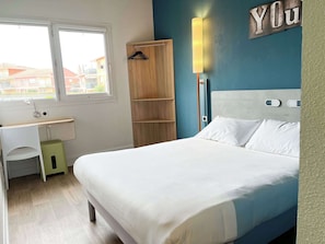 A bedroom with a bed, a desk, a wardrobe, and a wall-mounted light. - Ibis Budget Saint Christol Les Ales (Saint-Christol-Les-Ales)