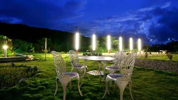 An outdoor seating area with white metal chairs and tables, surrounded by well-maintained grass and a stone border.