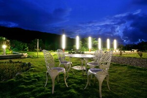 Un área de estar al aire libre con sillas y mesas de metal blanco, rodeada de césped cuidado y un borde de piedra.