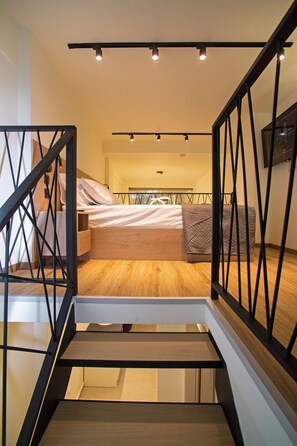 Un dormitorio moderno con suelo de madera, cama con ropa de cama blanca y marrón, una mesita de noche y un aparador. Hay una escalera con barandilla negra que lleva al dormitorio.