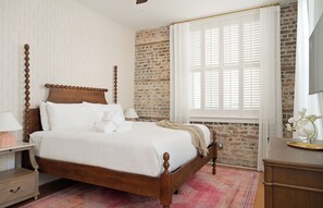A four-poster bed with white linens, a wooden nightstand with a lamp, a brick wall, and a window with white shutters. - The Conroy | Perfect Location | Historic Charm (Charleston)