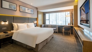 Une chambre d’hôtel avec un grand lit, un bureau et une chaise. Il y a deux tableaux encadrés au mur, une fenêtre avec des rideaux et un grand écran de télévision.