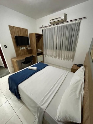 A hotel room with a bed, a desk, a chair, an air conditioning unit, and a television. - Pousada Serras De Goyaz (Goiânia)
