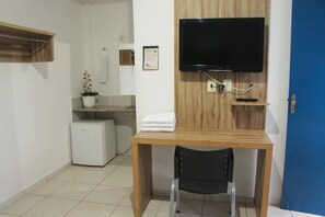 A small room with a wooden desk, a black chair, a television mounted on the wall, a small refrigerator, and a blue door.