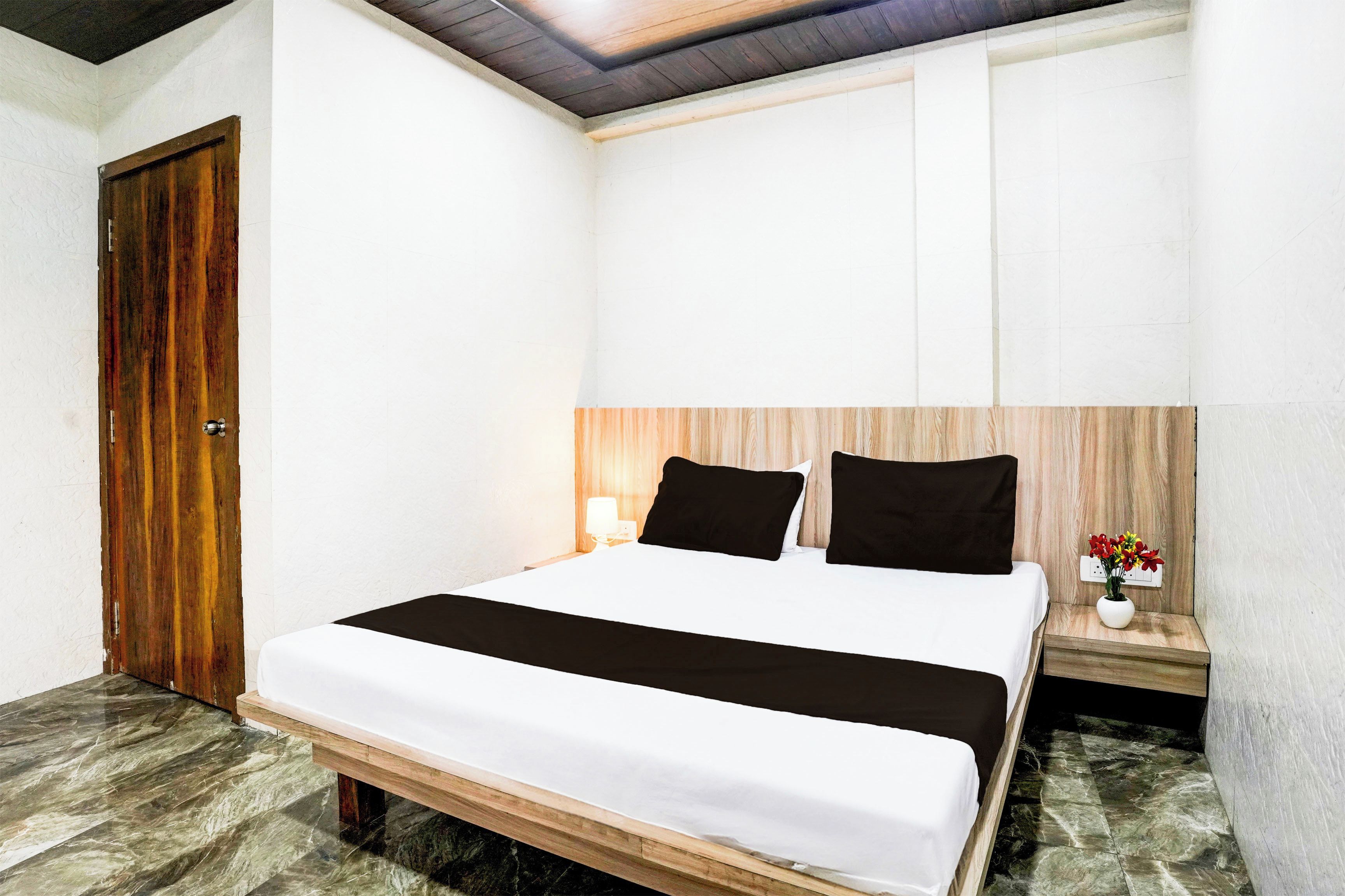 A hotel room with a bed, a wooden headboard, a nightstand with a lamp, and a small table with a vase of flowers.