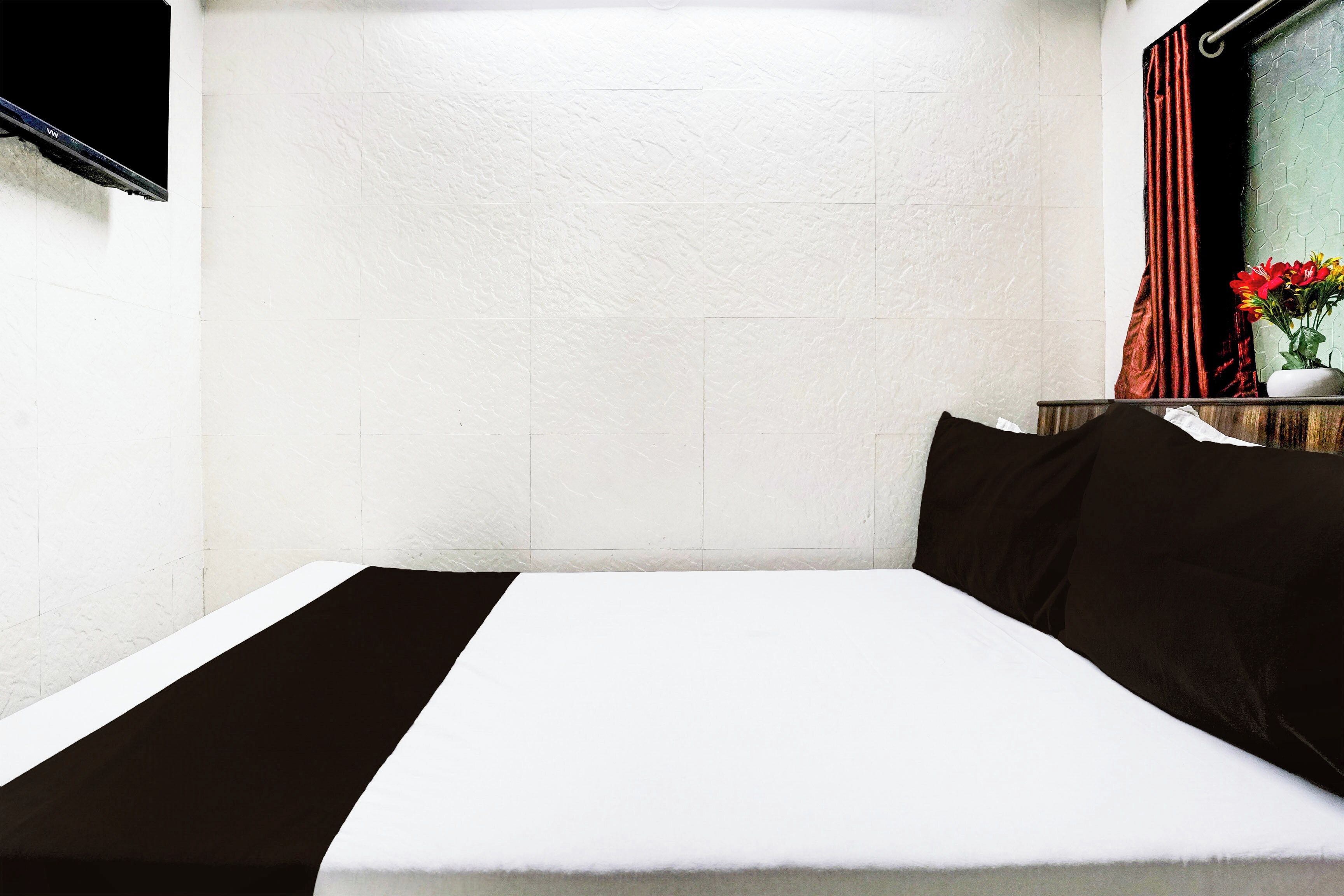 Una habitación de hotel con una cama, una ventana con cortinas y un florero con flores sobre una mesita de noche.