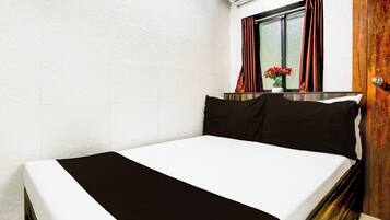 Una cama individual con una colcha blanco y negro en una habitación con paredes blancas, una puerta de madera y una ventana con cortinas rojas.