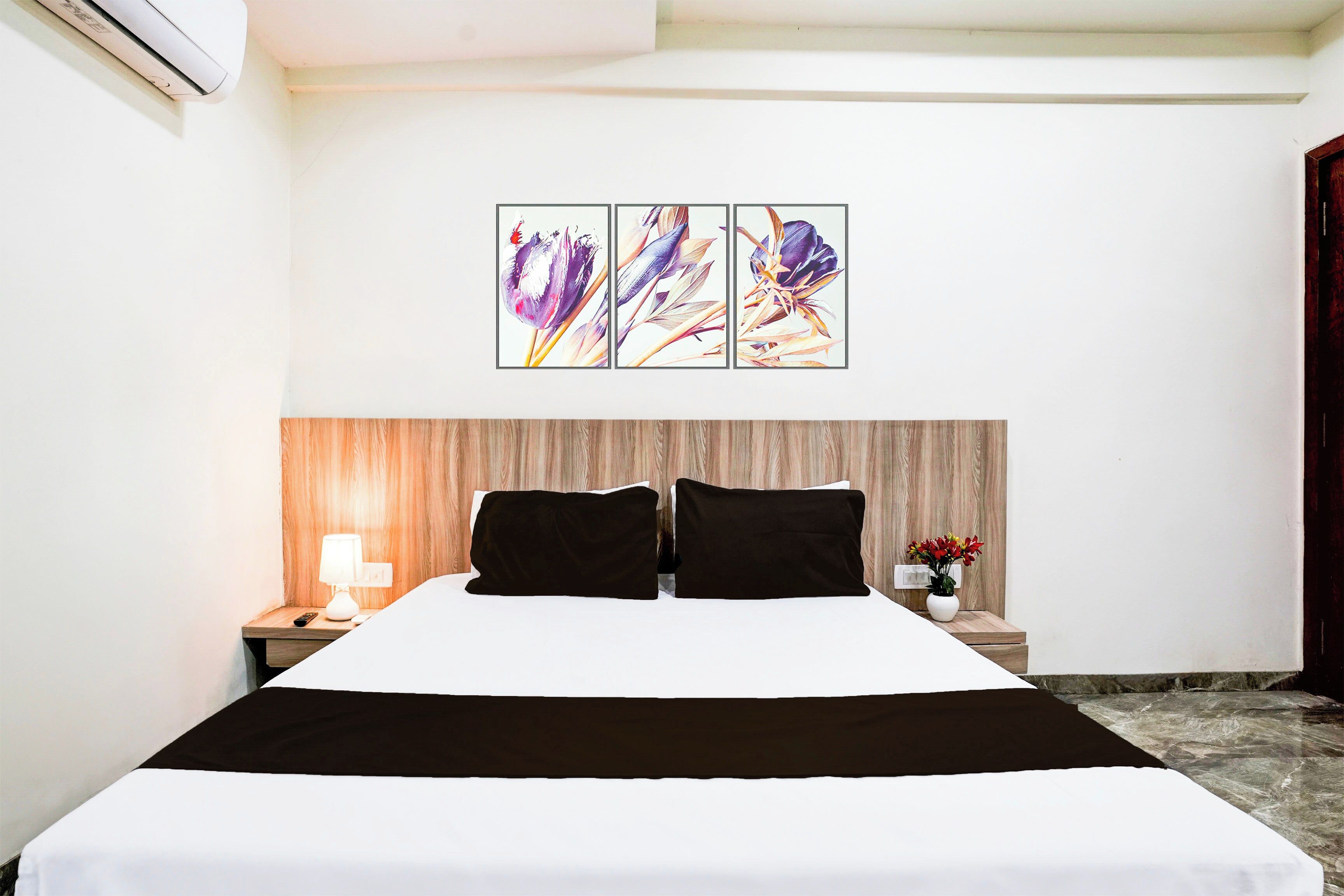 A hotel room with a bed, a nightstand with a lamp, a wall-mounted air conditioner, and a painting above the bed.