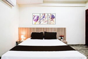 A hotel room with a bed, a nightstand with a lamp, a wall-mounted air conditioner, and a painting above the bed.